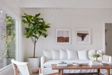 a boho living room with white furniture, a wooden coffee table, a small gallery wall and a tree plus a large window that allows a lot of natural light in