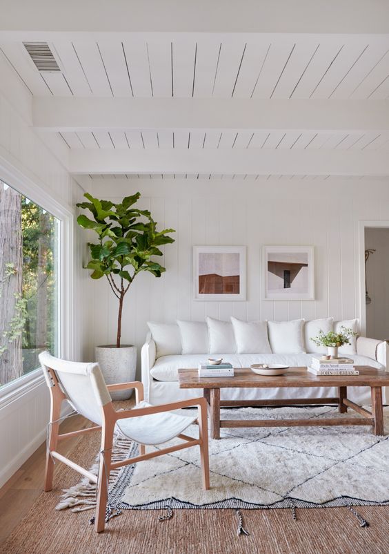 a boho living room with white furniture, a wooden coffee table, a small gallery wall and a tree plus a large window that allows a lot of natural light in
