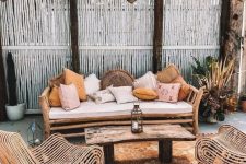 a boho tropical patio with a wooden bench and lots of pillows, catchy rattan chairs, a wooden table and a boho rug plus Moroccan lanterns