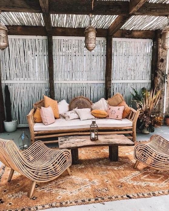 a boho tropical patio with a wooden bench and lots of pillows, catchy rattan chairs, a wooden table and a boho rug plus Moroccan lanterns