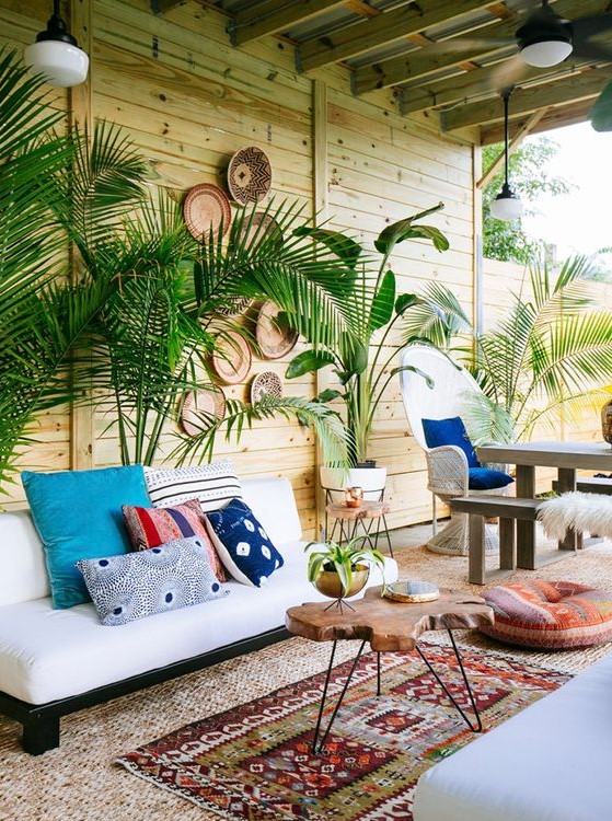 a boho tropical space with a wooden wall, a white sofa with colorful pillows, a living edge table and printed rugs