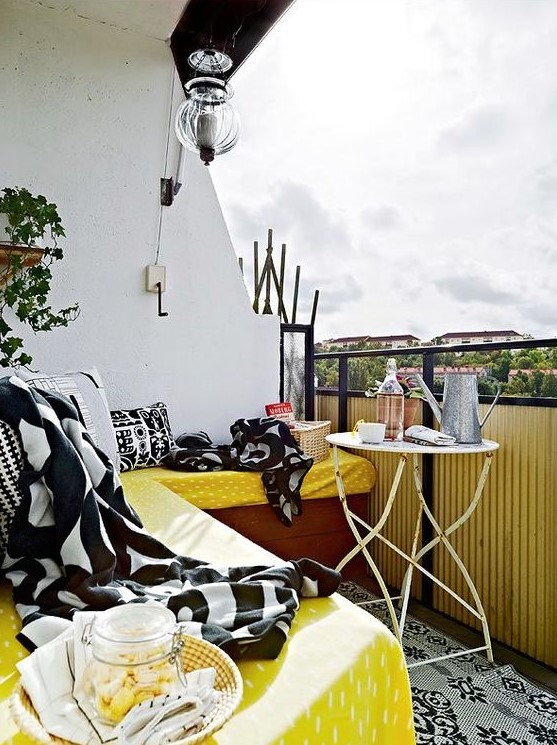 a bright summer balcony with a corner bench and yellow, black and white textiles, a shabby chic table and a potted plant