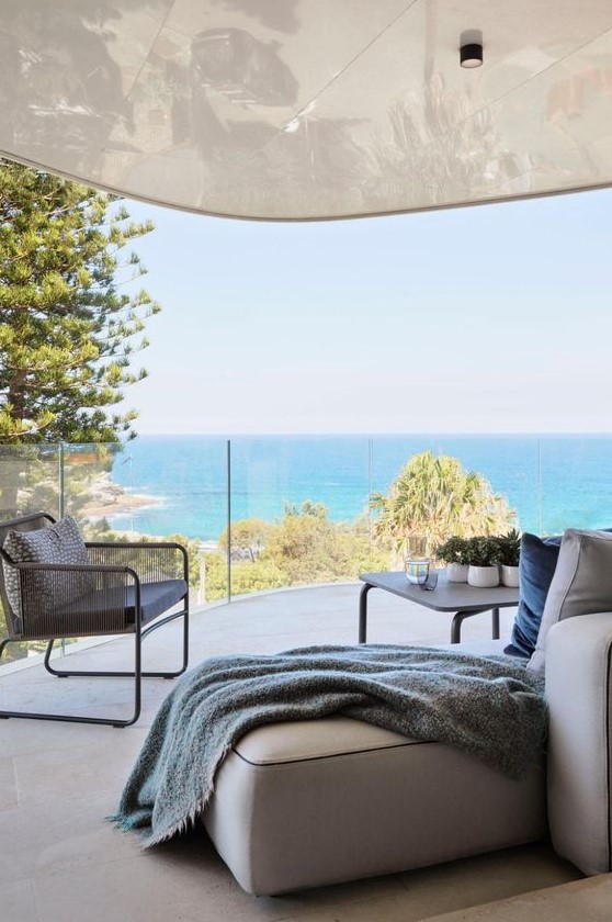 a coastal terrace with a neutral low sofa, a grey chair, a large coffee table and some potted greenery plus a sea view a coastal terrace with a neutral low sofa, a grey chair, a large coffee table and some potted greenery plus a sea view