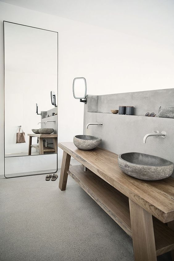 a contemporary bathroom with a large mirror in a sleek frame, a wooden vanity, carved stone sinks and a concrete wall