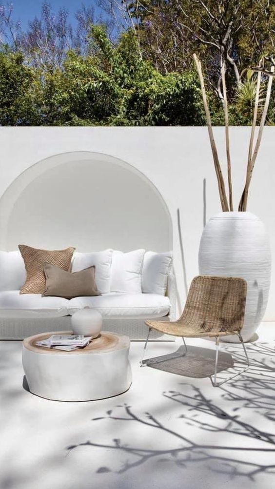 a contemporary neutral terrace with a white wicker sofa in the niche, white pillows, a neutral chair and a whitewashed tree stump table a contemporary neutral terrace with a white wicker sofa in the niche, white pillows, a neutral chair and a whitewashed tree stump table