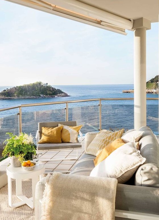 a contemporary seaside terrace with neutral furniture, pillows, a small coffee table and a neutral rug and a lovely sea view a contemporary seaside terrace with neutral furniture, pillows, a small coffee table and a neutral rug and a lovely sea view
