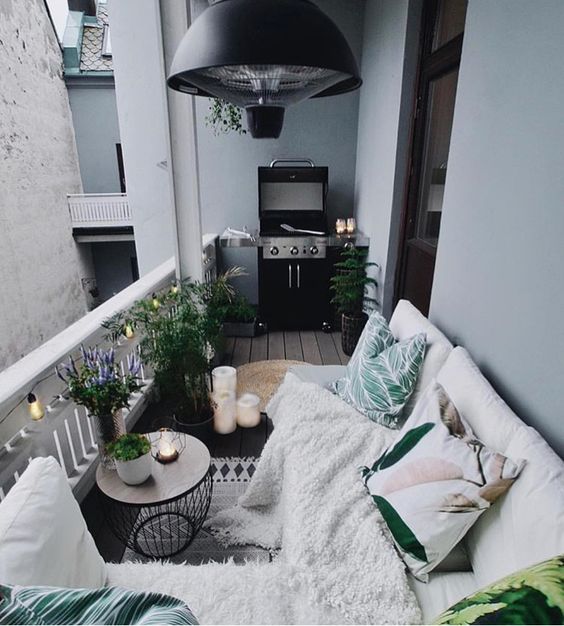 a cozy boho terrace with a white sofa, bright pillows and a blanket, a side table with potted greenery and blooms plus a grill