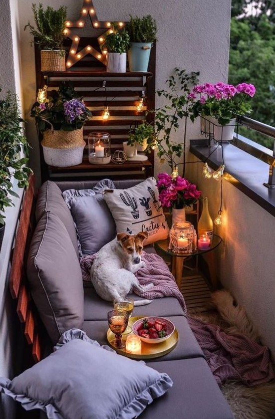 a cozy small balcony with a pallet wall and sofa, some potted blooms and greenery and candle lanterns