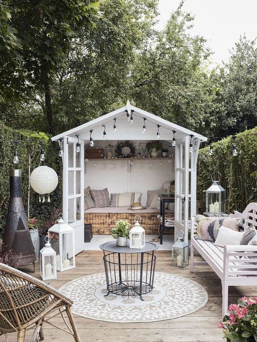 a cute boho outdoor dining space a cozy small terrace with a hearth, a white bench with lots of pillows, a coffee table, a rattan chait, lots of lanterns and a small shed with a daybed