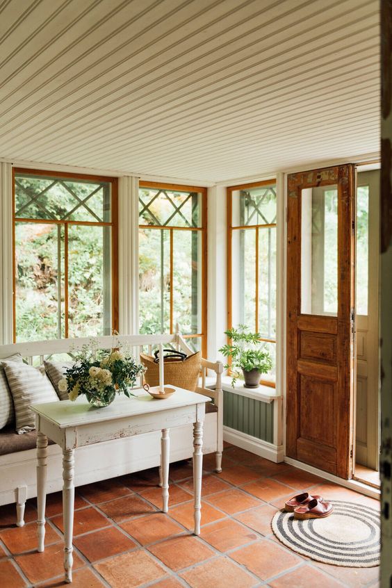 a cozy vintage Scandinavian sunroom with a tiled floor, vintage white furniture, potted blooms and greenery and printed pillows