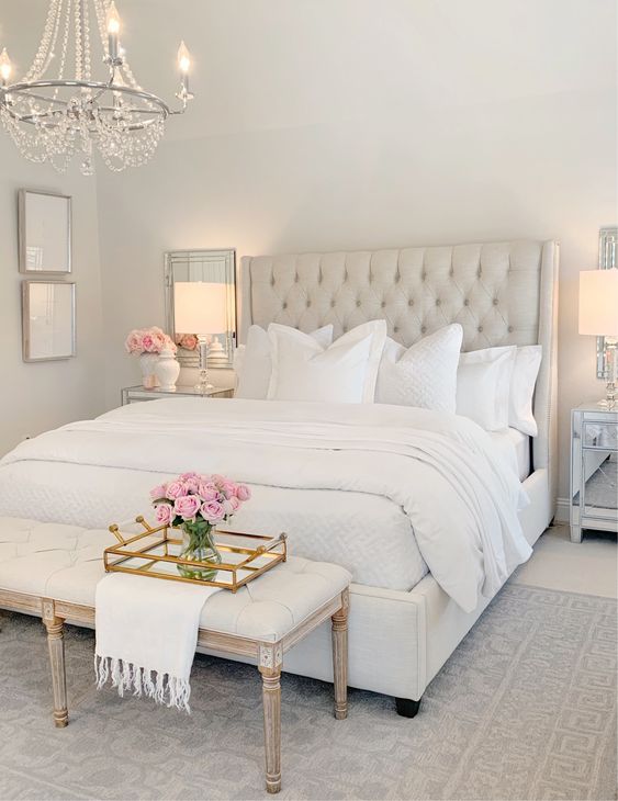 a glam and vintage neutral bedroom with a creay bed, a crystal chandelier, a neutral bench, mirrors and artworks