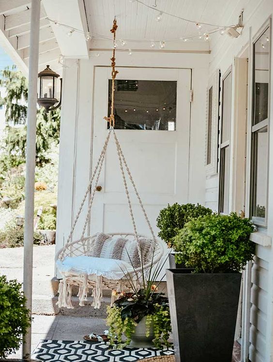 a hygge porch with a neutral pendant chair with pillows, potted greenery, string lights and layered rugs is cozy