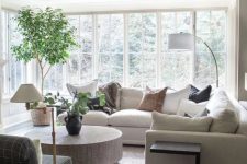 a light-filled neutral living room with a large sectional, a floor lamp, a round coffee table, a graphite grey chair and some greenery