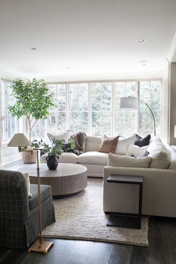 a light-filled neutral living room with a large sectional, a floor lamp, a round coffee table, a graphite grey chair and some greenery