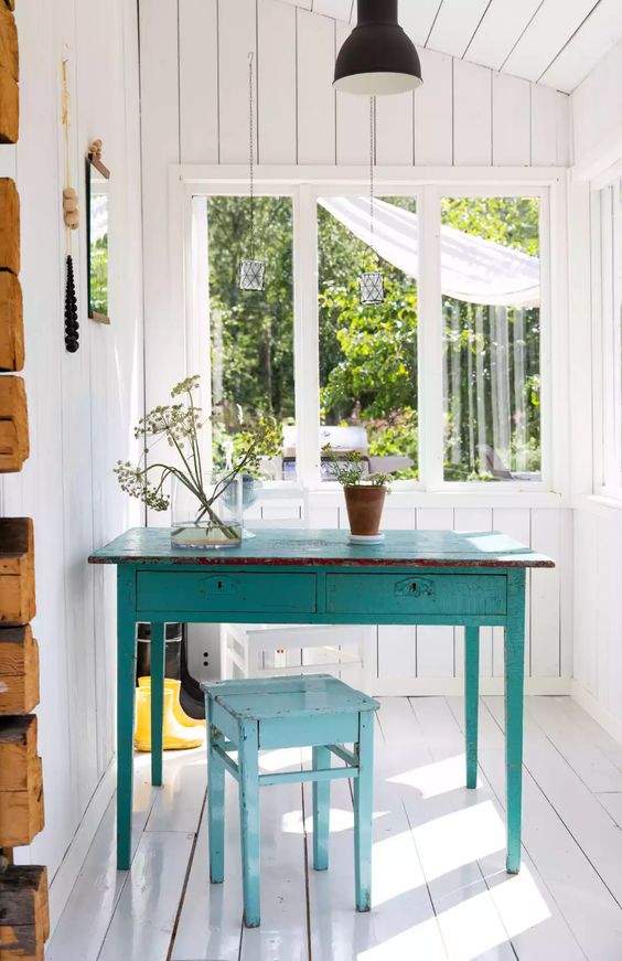 a little white sunroom with planked walls and a floor, a blue table and a stool, a black pendant lamp and potted greenery is cool