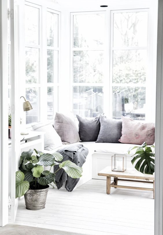 a lovely Nordic sunroom in white, with a built in sectional sofa with pastel pillows, potted plants and a low wooden coffee table