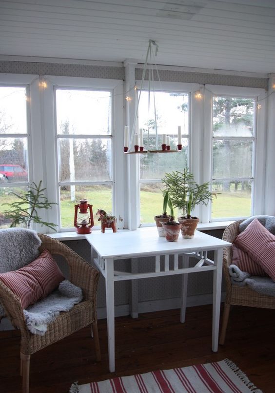 a lovely Scandinavian sunroom with a planked ceiling, woven chairs, a white table, some faux fur and a chandelier with candles