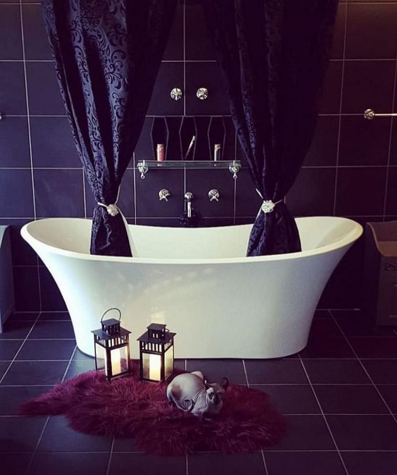 a modern Gothic bathroom with black tiles, a white tub with printed black curtains, a glass shelf and coffin-shaped niches