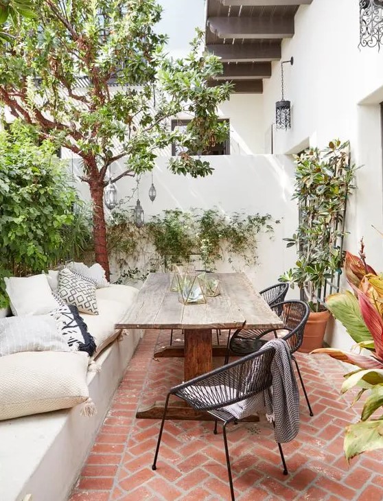 a modern country patio with a birck floor, a built in bench, a vintage wooden table, some wicker chairs and potted plants and trees a modern country patio with a birck floor, a built-in bench, a vintage wooden table, some wicker chairs and potted plants and trees
