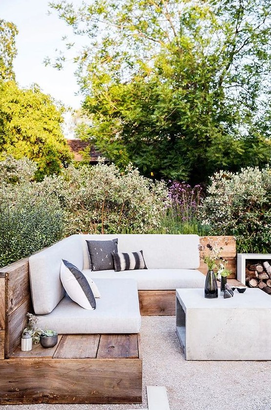 a modern farmhouse outdoor living room with a large built-in corner sofa, a concrete coffee table, printed pillows, a firewood storage unit