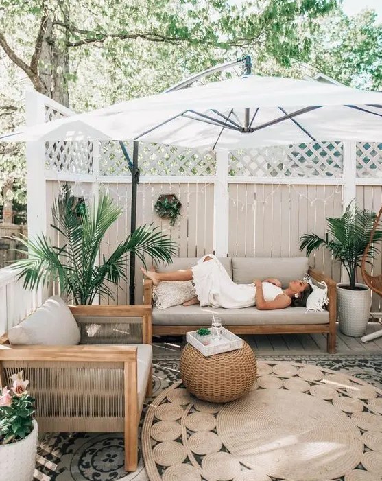 a modern tropical patio with wooden furniture, a woven side table, potted plants, layered rugs and an umbrella over the space
