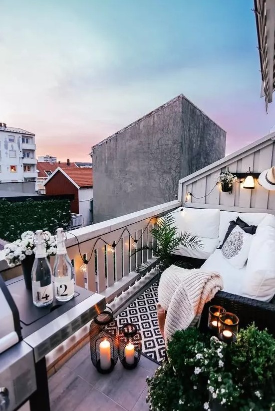 a monochromatic small balcony with black wicker furniture, candle lanterns, potted greenery and blooms and a grill