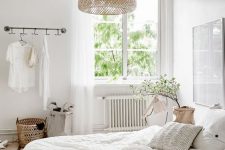 a neutral Scandinavian bedroom with white furniture, a pendant woven lamp, neutral bedding and some greenery