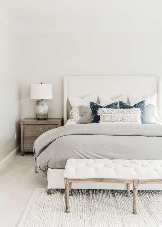 a neutral bedroom with a creamy bed, a white upholstered bench, wooden nightstands and printed pillows