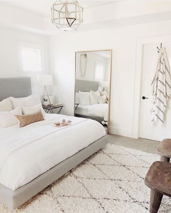 a neutral bedroom with a grey upholstered bed, an oversized mirror, a pendant lamp and printed pillows
