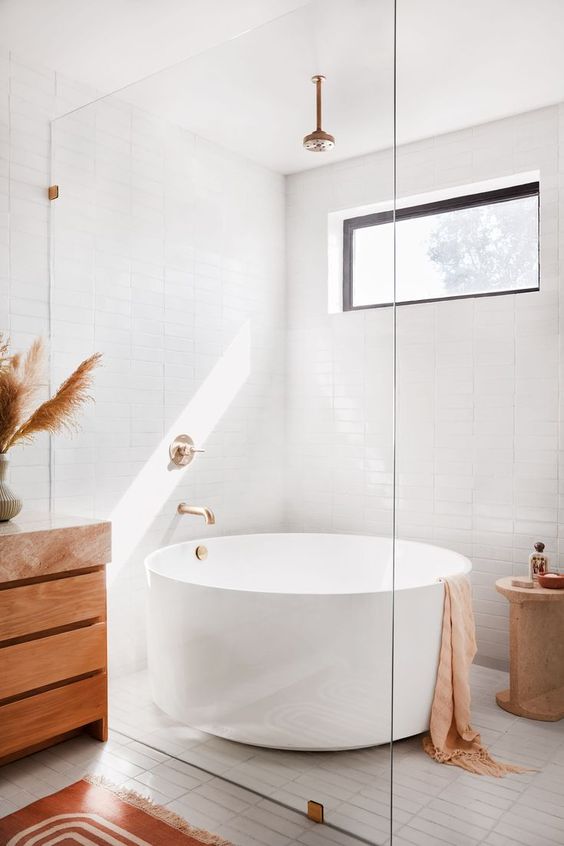 a neutral boho bathroom with a window, a round bathtub, a wooden vanity, pampas grass, a wooden side table and tan textiles