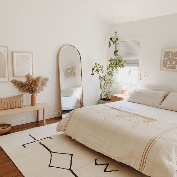 a neutral boho bedroom with a bed, a wooden bench, a curved mirror, potted plants and artworks