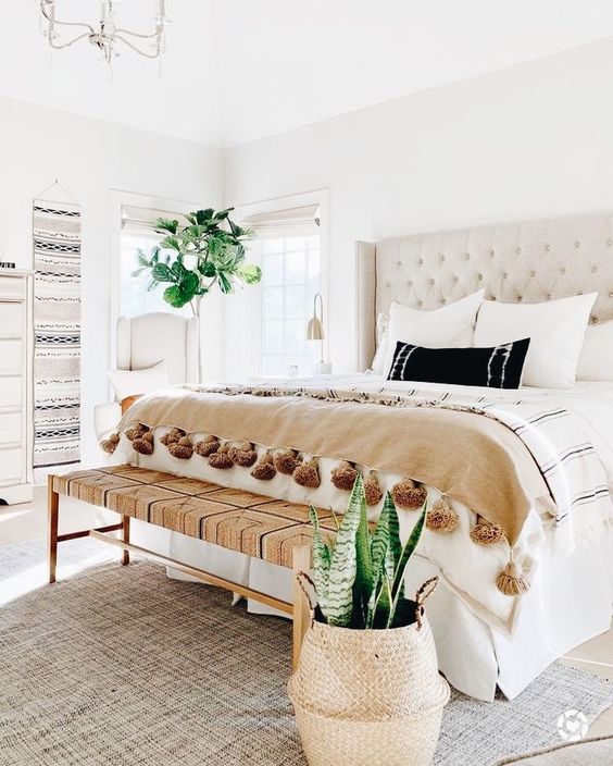 a neutral boho bedroom with a creamy bed, a woven bench, boho hangings, statement plants and tassel blankets