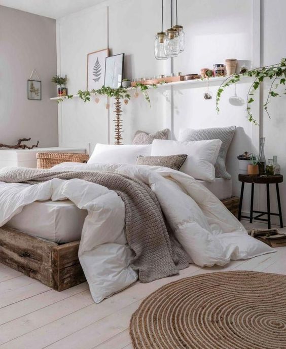 a neutral boho bedroom with a rough wooden bed, a jute rug, an open shelf, pendant lamps and greenery