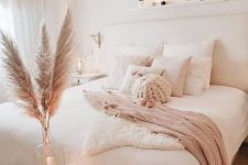 a neutral boho bedroom with white furniture, a woven lamp, pampas grass and candles