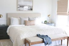 a neutral coastal bedroom with an upholstered bed, woven shades and a lamp, pillows and a long bench