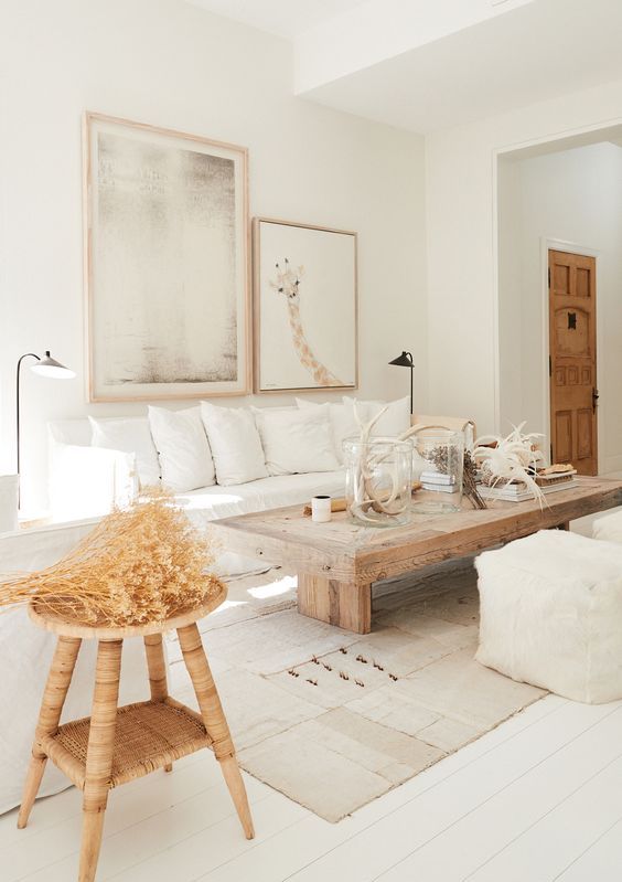 a neutral living room ith white furniture, a wooden table and stool, layered rugs, a gallery wall and some decor on the table