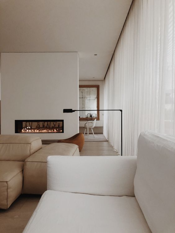a neutral minimalist living room with double sided fireplace, a tan leather sofa, a white one and an orcher chair by the fireplace