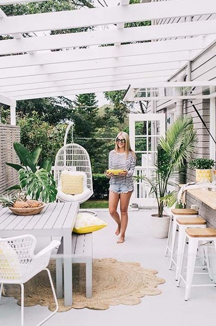 a neutral modern tropical terrace with white furniture, metal and wood stools, a jute rug and a hanging chair