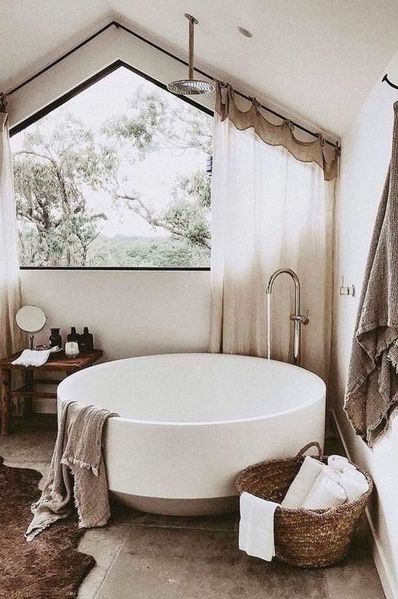 a neutral sabi-sabi bathroom with tiles on the floor, a round tub, a basket with towels, a wooden stool and curtains on the window