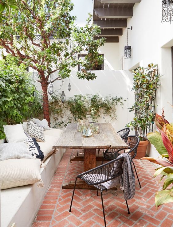 a neutral terrace with a built-in bench with lots of pillows, a stained table and black chairs and lots of greenery around