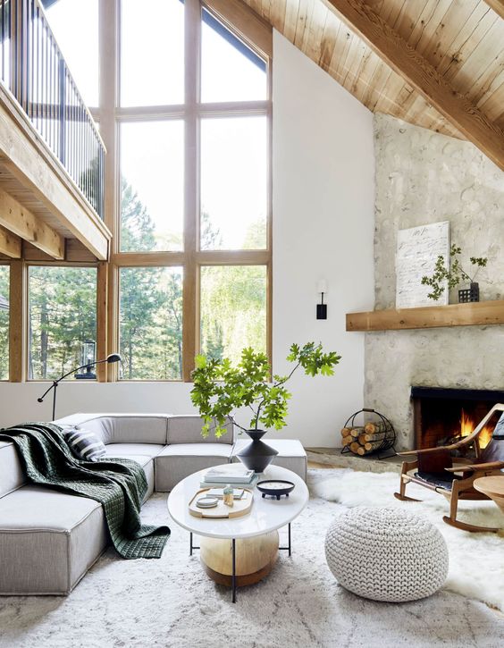 a peaceful neutral living room with double height ceilings, a fireplace clad with stone, a grey sectional, an oval coffee table and a knit pouf