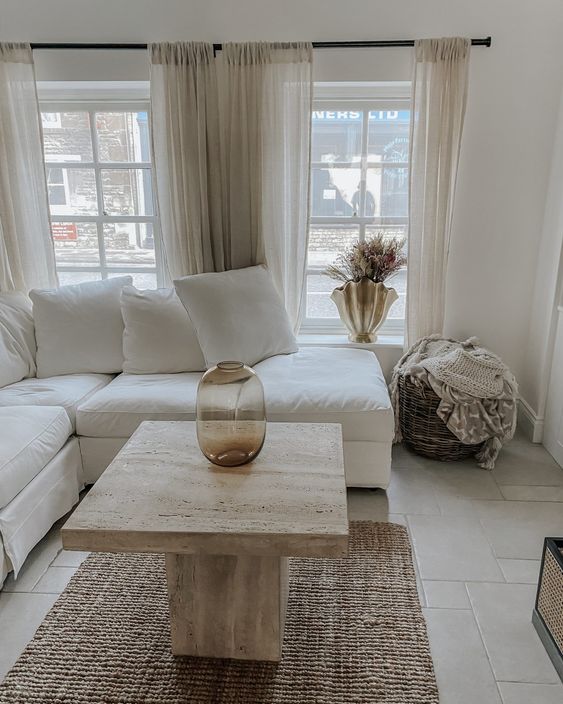 a pretty neutral living room with a white sectional, a stone coffee table, a woven rug, sheer curtains and a basket for blankets