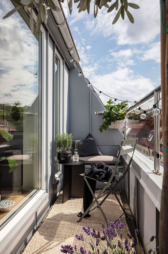 a pretty small Nordic balcony with a smallbench, a folding chair, a black table and a jute rug plus potted greenery