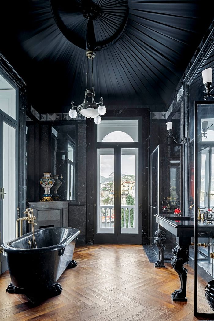 a quirky Gothic bathroom in black, with an attic textural ceiling, a black stone tub and black stone furniture with paws, with a fireplace and a large mirror
