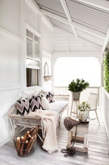 a relaxed Scandinavian porch with a white shabby daybed and white chairs and tables, firewood, greenery and cozy blankets and pillows