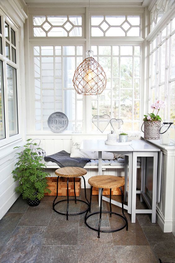 a small Scandi sunroom with a built-in bench, a white table, stained stools, potted greenery and a woven pendant lamp