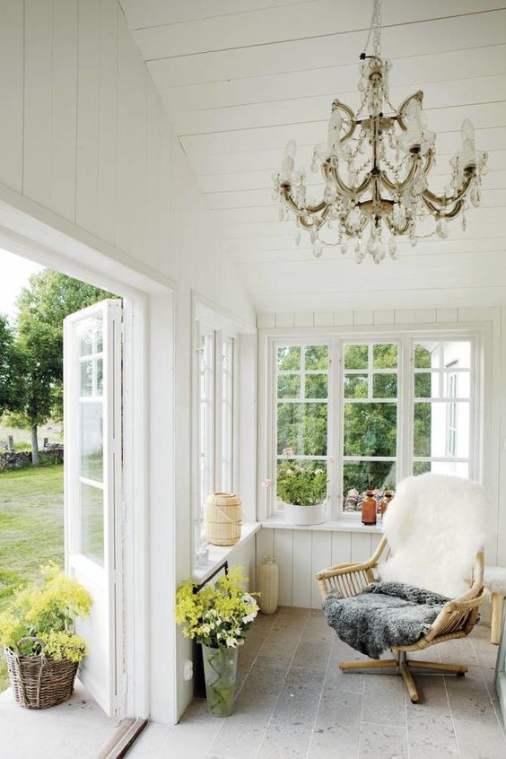 a small Scandi sunroom with a rattan chair with faux fur, a vintage crystal chandelier, potted blooms and greenery can be opened to outdoors