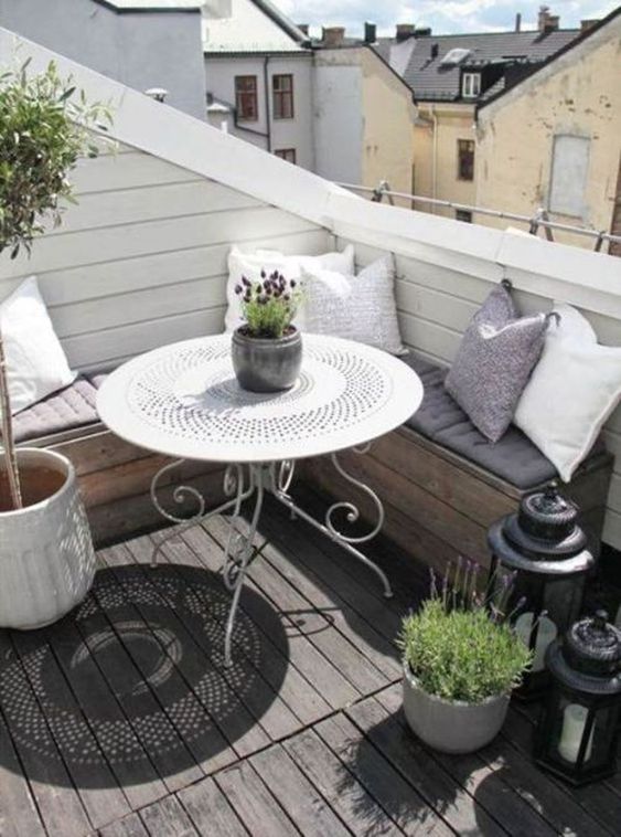 a small Scandinavian terrace with a built-in L-shaped bench, a forged table, potted greenery and blooms plus candle lanterns