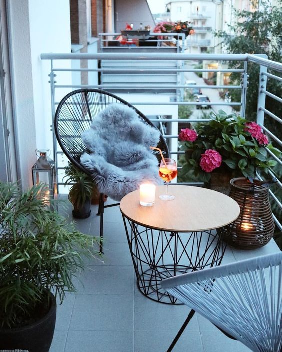 a small and chic balcony with wicker chairs, a round coffee table, potted blooms and plants and candle lanterns