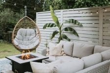 a small and cool farmhouse patio with a neutral wicker corner sofa, a fire pit, a pendant egg chair and a potted plant